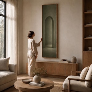 Puede incluir: Una pieza de arte mural rectangular y alta que presenta una pintura de color verde oliva apagado de un arco de piedra. La obra está enmarcada en un borde de madera clara y delgada, montada verticalmente sobre una pared neutra y un mueble.