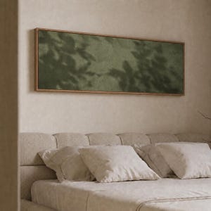 May include: A rectangular wall art piece featuring a textured olive green surface with dark, leafy shadow patterns cast across it. The artwork is enclosed in a thin, light-toned wooden frame, mounted horizontally above a neutral-colored bed.