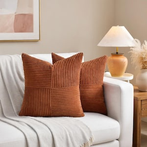May include: Two rust-colored corduroy throw pillows with a geometric design rest on a white sofa. The pillows have a textured, ribbed appearance and a square shape. The sofa is covered with a white blanket.