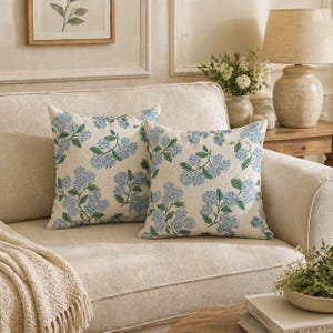 Puede incluir: Dos cojines cuadrados con fondo crema y un estampado floral de hortensias azules. Los cojines están colocados sobre un sofá beige, creando un ambiente acogedor y agradable. Añaden un toque de decoración inspirada en la naturaleza.