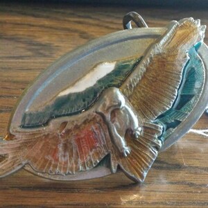 Pode incluir: Fivela de cinto de metal vintage com um design de águia. A fivela oval apresenta um relevo detalhado de uma águia com as asas abertas, pintada em tons de ouro, vermelho e azul. O fundo inclui uma cena de montanha. Uma pequena etiqueta está anexada.