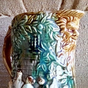 May include: A vintage ceramic pitcher with a detailed relief design. The pitcher features a handle and a base with a decorative border. The body of the pitcher depicts a scene with figures, trees, and a building, in shades of green, blue, brown, and beige.