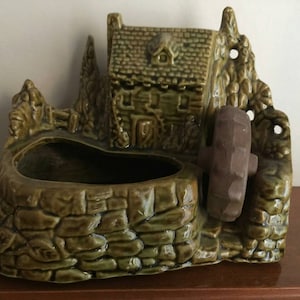 May include: A green ceramic planter shaped like a cottage with a water wheel. The planter has a textured, stone-like finish. The cottage features a small door and windows, and the water wheel is a dark brown colour.