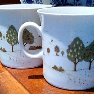May include: Two white ceramic mugs with a landscape design featuring trees with green foliage and red fruit. The mugs have a curved handle and a slightly flared rim. The design is in shades of brown and green.