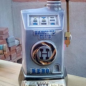 May include: A vintage ceramic decanter shaped like a slot machine, with the words "HAROLDS CLUB RENO" and "BEAM 100" visible. The decanter has a pull lever and a clear glass stopper. The base reads "Kentucky Straight Bourbon Whiskey".