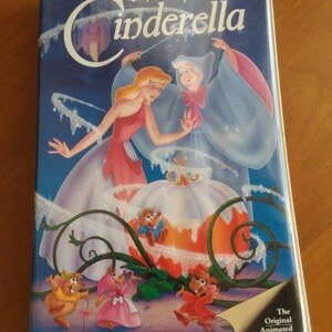 Puede incluir: Una cinta de VHS del clásico animado de Disney, Cenicienta. La portada presenta a Cenicienta con un vestido rosa, el Hada Madrina y una carroza de calabaza. El título está en escritura blanca sobre fondo azul.