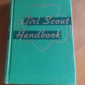 Op de afbeelding: Een groene Girl Scout Handbook met zilveren letters. Het boek heeft een rode omtrek van het Girl Scout-logo. Het boek is gesloten en ligt op een houten oppervlak.