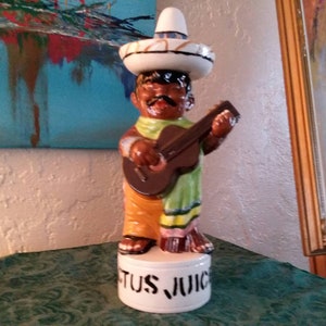 May include: A ceramic figurine of a mariachi musician playing a guitar. The figure wears a sombrero, a green and yellow serape, and orange pants. The base of the figurine reads "TUS JUICE".