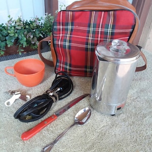 May include: A vintage picnic set featuring a red plaid backpack with brown leather accents, a stainless steel coffee pot, an orange cup, a knife, a spoon, keys, and a black electrical cord. The set is arranged on a textured surface.