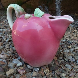 May include: A pink ceramic pitcher shaped like an apple, with a white handle and a green leaf accent. The pitcher has a glossy finish and is sitting on a bed of small rocks.