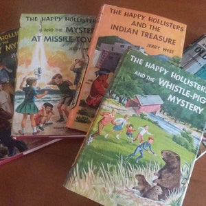 May include: A collection of vintage children's books titled "The Happy Hollisters." The book covers feature colorful illustrations and titles like "The Cowboy Mystery," "The Mystery at Missile Town," and "The Indian Treasure."