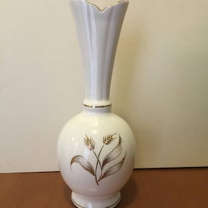 May include: White ceramic vase with a bulbous base and a flared, scalloped top. The vase features a gold wheat stalk design and gold trim. A decorative piece for displaying flowers or as a standalone accent.