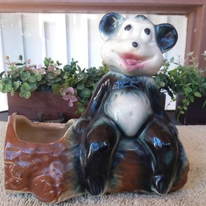 May include: A ceramic monkey planter with a brown and black body, blue ears, and a smiling face. The planter has a rectangular opening for plants. The monkey is sitting on a brown log-shaped base.