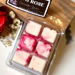 Puede incluir: Un paquete de plástico transparente que contiene seis ceras perfumadas cuadradas. Las ceras son blancas con vetas rosas. Una etiqueta en la parte superior izquierda dice "Dubai Rose" y "Chocolate Dubai". El paquete está sobre una superficie de madera.