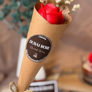 Puede incluir: Una rosa roja en un cono de papel marrón, atada con hilo. El cono tiene una etiqueta circular con el texto "Dubai Rose Chocolate Dubai" y el nombre "Chloeta". Se ven pequeñas flores blancas.