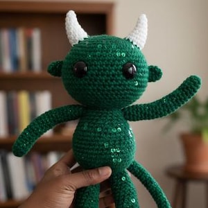 Puede incluir: Muñeca verde de ganchillo hecha a mano con cuernos y detalles blancos. La muñeca tiene ojos negros y está adornada con lentejuelas. La muñeca está sujeta en la mano de una persona. El fondo incluye una estantería y una planta en maceta.