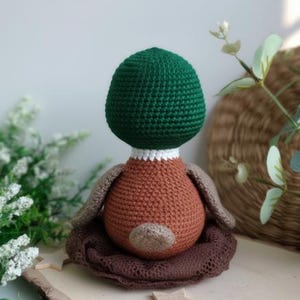 May include: A handmade crochet duckling doll with a green head, brown body, and tan wings. The duckling sits in a brown crocheted nest on a wooden surface. A charming, soft toy for children or as a decorative item.