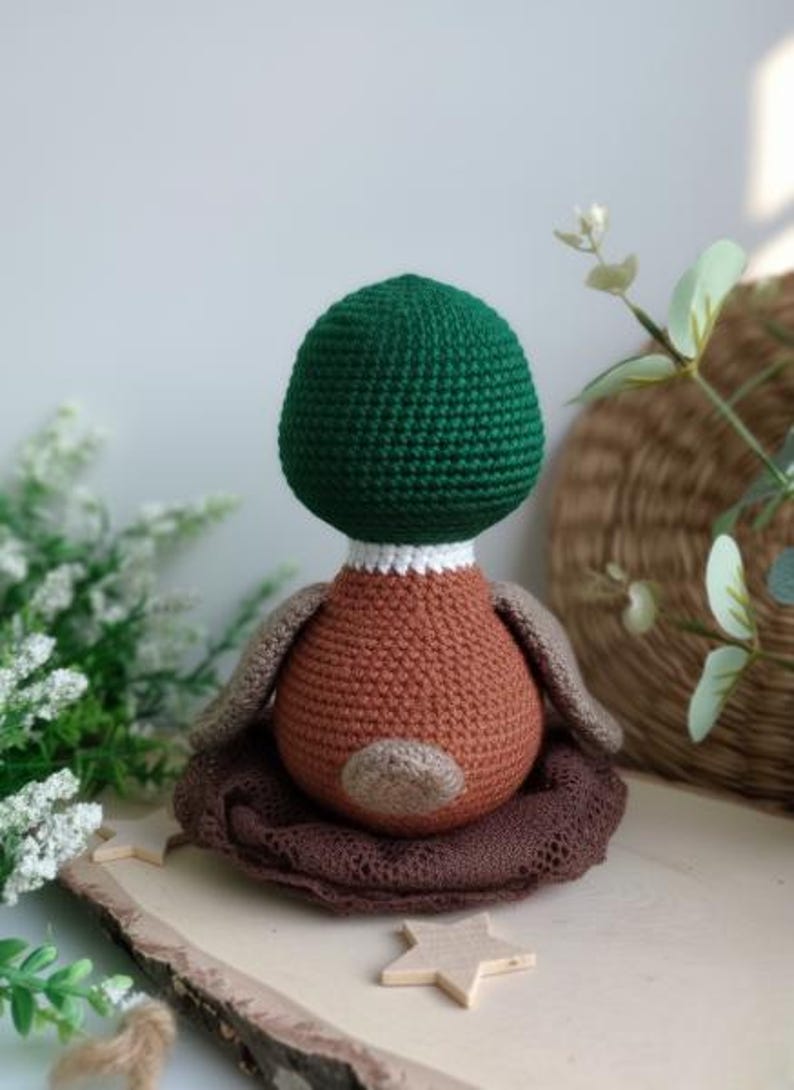 May include: A handmade crochet duckling doll with a green head, brown body, and tan wings. The duckling sits in a brown crocheted nest on a wooden surface. A charming, soft toy for children or as a decorative item.