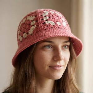 May include: A crochet bucket hat featuring a dusty rose pink and cream white granny square pattern. The hat is constructed with a rounded crown and a downward-sloping brim, showcasing intricate yarn stitching throughout the entire design.