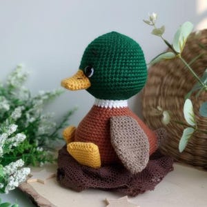 May include: A handmade crochet duckling with a green head, brown body, and yellow feet, sitting on a brown nest. The duck has a yellow beak and white collar. The duck is a charming, handcrafted item.