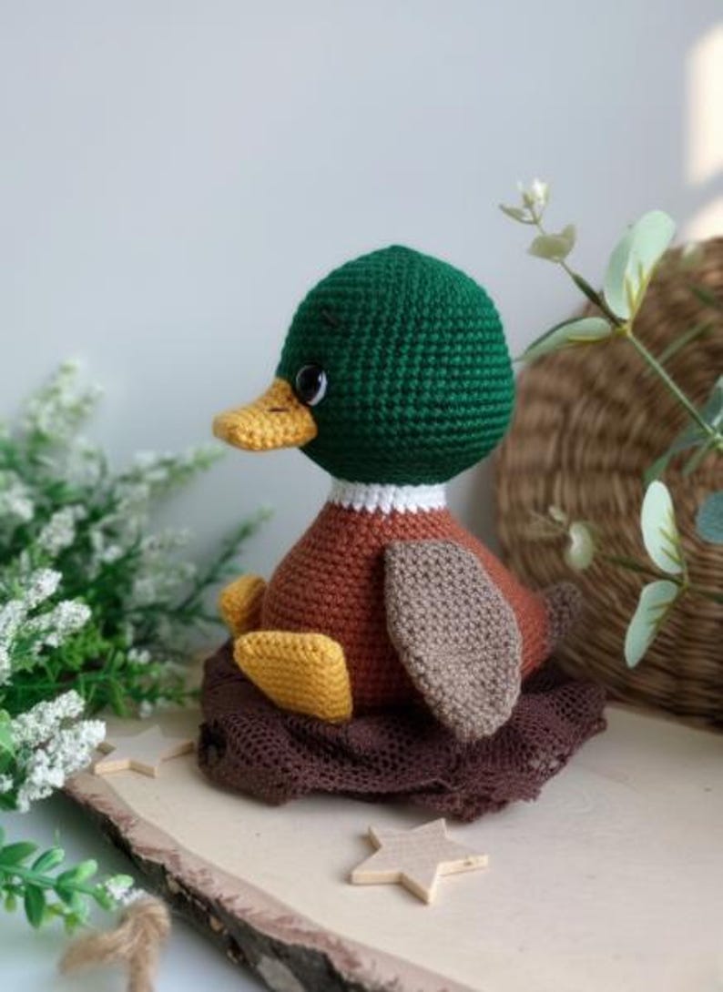 May include: A handmade crochet duckling with a green head, brown body, and yellow feet, sitting on a brown nest. The duck has a yellow beak and white collar. The duck is a charming, handcrafted item.