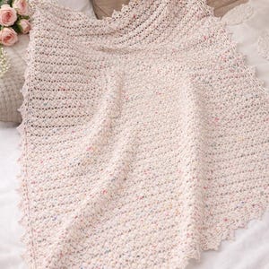 Puede incluir: Una manta crema tejida a mano con un borde festoneado. La manta tiene una apariencia texturizada con motas de color. La manta está doblada, mostrando el intrincado trabajo de puntadas y el diseño. La manta mide aproximadamente 152 cm de ancho y 178 cm de largo.