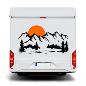 Pode incluir: Parte traseira de uma autocaravana branca com um decalque preto de montanha e floresta. Um sol laranja está centrado acima da cordilheira. O decalque apresenta dois pinheiros em cada lado da floresta.