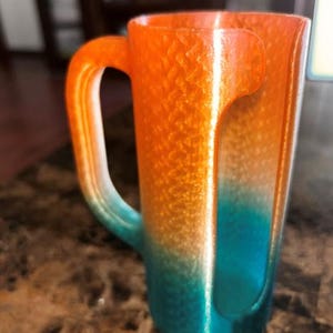 May include: A gradient-colored mug with a unique handle design. The mug transitions from orange to teal, with a textured surface. The handle is a continuation of the mug's body, creating a seamless look. The mug is likely made of a durable material.