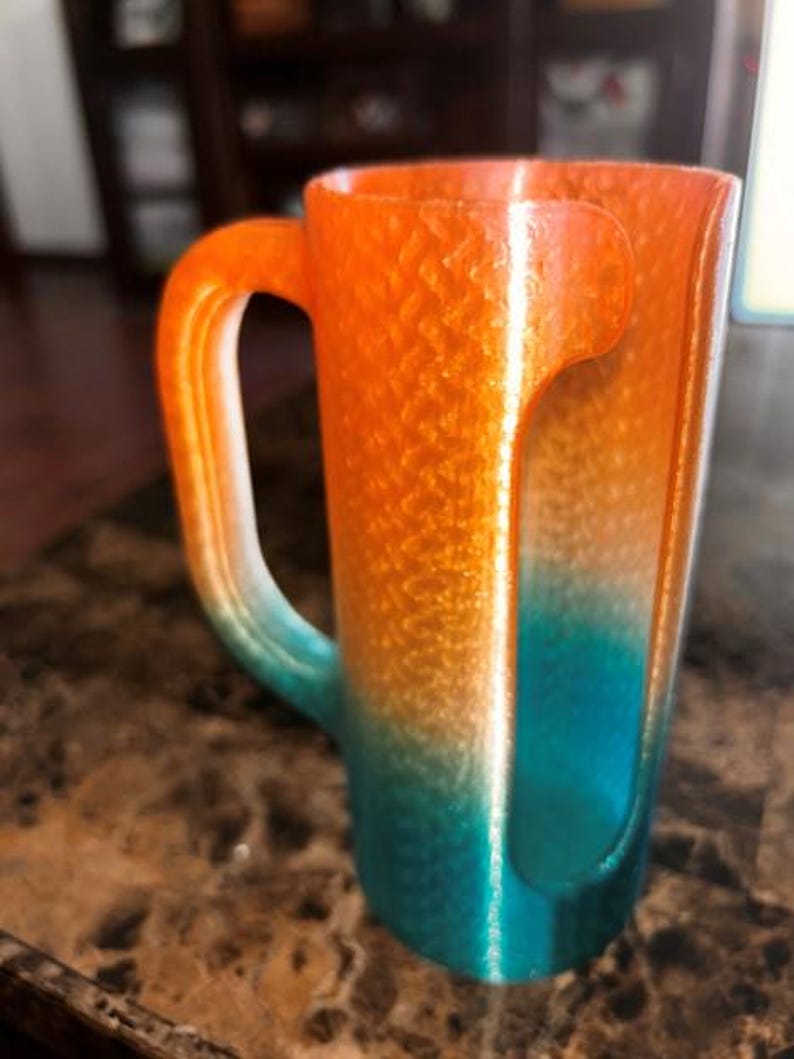 May include: A gradient-colored mug with a unique handle design. The mug transitions from orange to teal, with a textured surface. The handle is a continuation of the mug's body, creating a seamless look. The mug is likely made of a durable material.