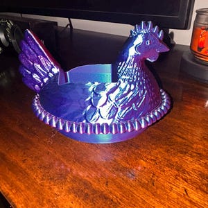 May include: A decorative hen-shaped container in iridescent blue and purple hues. The hen is perched on a nest-like base with a beaded rim. The container is likely used for storage or display.