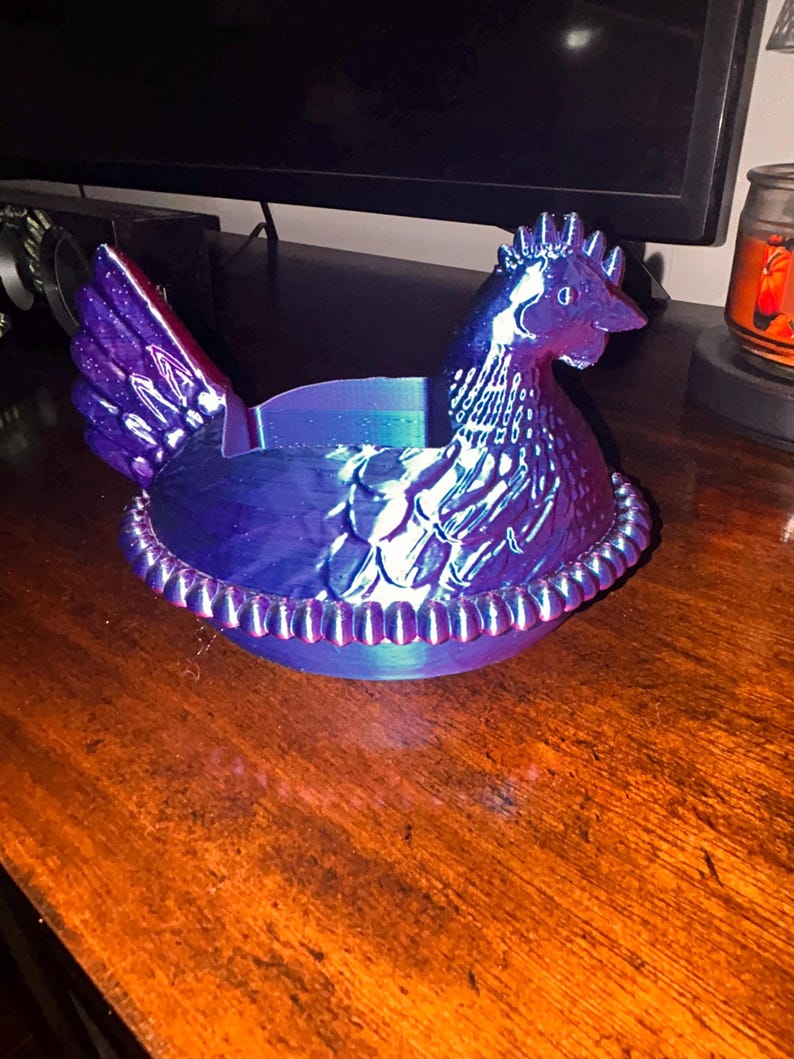 May include: A decorative hen-shaped container in iridescent blue and purple hues. The hen is perched on a nest-like base with a beaded rim. The container is likely used for storage or display.