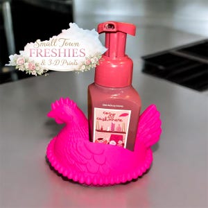 May include: A pink soap dispenser with a matching pump sits inside a bright pink chicken-shaped holder. The soap bottle has a label that reads "cozy cashmere." The image also includes a sign that says "Small Town Freshies & 3-D Prints."