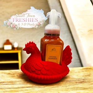 May include: A red chicken-shaped holder cradles a bottle of Kitchen Mandarin hand soap. The soap bottle is orange with a yellow label. The holder is made of a solid red material and has a detailed design. The image includes the text "Small Town Freshies & 3-D Prints".