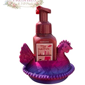 May include: A burgundy soap dispenser with a red pump sits in a 3D-printed hen-shaped holder. The holder is a gradient of purple and pink. The soap bottle has a label that reads "cozy cashmere". The image also includes the text "Small Town Freshies & 3-D Prints".