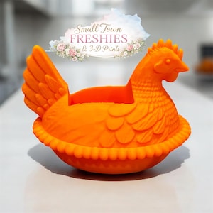 May include: Bright orange hen-shaped container with a detailed design. The hen has a textured body, a comb, and a beak. The container has a ruffled edge and a space for storage. The text "Small Town Freshies & 3-D Prints" is above the hen.