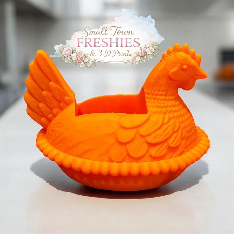 May include: Bright orange hen-shaped container with a detailed design. The hen has a textured body, a comb, and a beak. The container has a ruffled edge and a space for storage. The text "Small Town Freshies & 3-D Prints" is above the hen.