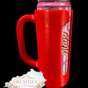 May include: A bright red 3D-printed can holder with a handle, designed to hold a standard-sized beverage can. The can is pink and white with text. The holder has a cutout to display the can's label. The logo for Small Town Freshies & 3-D Prints is visible.