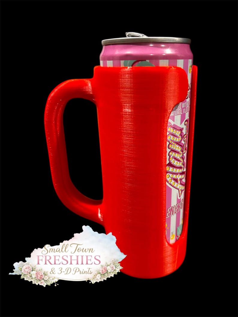 May include: A bright red 3D-printed can holder with a handle, designed to hold a standard-sized beverage can. The can is pink and white with text. The holder has a cutout to display the can's label. The logo for Small Town Freshies & 3-D Prints is visible.