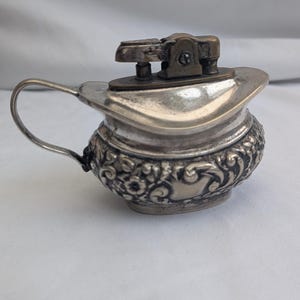 May include: A vintage silver-toned lighter with a unique pitcher-like design. The lighter features a decorative floral pattern around the base and a handle. The top portion has a hinged mechanism. The lighter is likely made of metal.