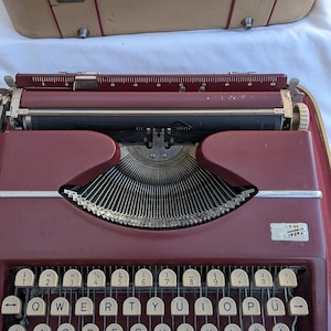 May include: A burgundy vintage typewriter with off-white keys, in its open burgundy carrying case. The keys are arranged in a QWERTY layout. The brand name "Tippa" is visible on the front. The typewriter's case is beige.