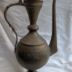 May include: An antique bronze-coloured metal pitcher with detailed floral and animal engravings. It has a long, curved spout, a handle, and a conical lid. The pitcher stands on a round base, displaying a vintage aesthetic.