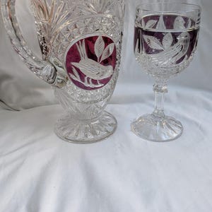 May include: A clear glass jug and wine glass set. The jug has a handle and a wide base. Both pieces feature a bird and leaf design in a burgundy oval. The glass has a stem and a wide bowl.