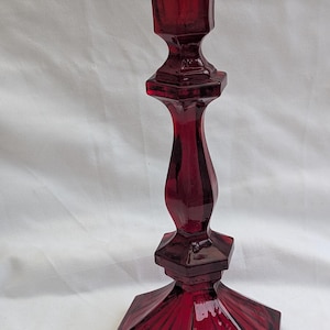 May include: A deep red glass candlestick holder with a geometric design. The candlestick has a hexagonal base, a decorative centre, and a flared top. The glass is transparent. The candlestick is a decorative item, suitable for holding a candle.