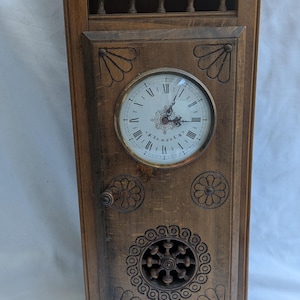 Puede incluir: Un reloj antiguo de madera con acabado marrón claro. La esfera del reloj es blanca con números romanos negros. El reloj tiene tallas decorativas y una pequeña puerta con un asa redonda. La parte superior tiene un diseño tallado decorativo.