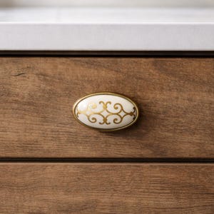 May include: Oval cabinet pull with a white ceramic center and gold filigree design. The pull has a gold-colored metal frame and is mounted on a wooden cabinet door. The cabinet door is a medium brown color.