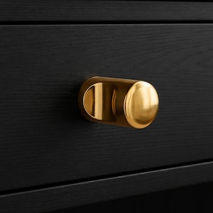 May include: Close-up of a gold-colored cylindrical drawer pull mounted on a black wooden surface. The pull has a rounded end and a curved, recessed base, providing a modern aesthetic. The wood grain is visible.
