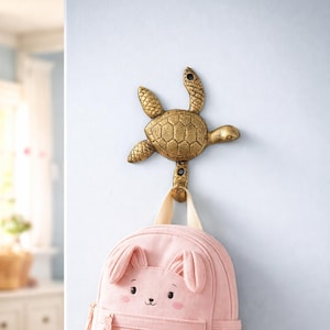 May include: A gold-colored metal turtle wall hook with a pink backpack hanging from it. The turtle hook is mounted on a light blue wall. The backpack has a bunny face design.