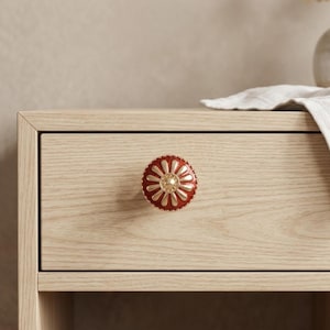 May include: A red and gold decorative drawer knob with a floral design. The knob is mounted on a light wood drawer. The knob has a gold center and a red outer ring with gold accents.