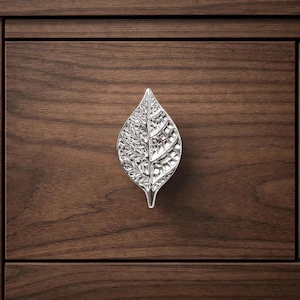 May include: A silver leaf-shaped cabinet pull is mounted on a dark brown wooden drawer. The pull has intricate detailing, resembling the veins of a leaf. The wood grain is visible in the background, creating a natural contrast.