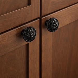 May include: Close-up of two black cabinet knobs with a floral design. The knobs are round with a detailed, textured surface and are mounted on a wooden cabinet with a warm brown finish. The knobs add a decorative touch.